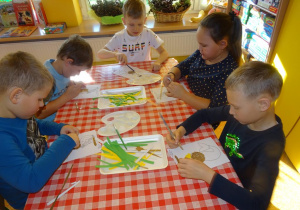 Piątka dzieci siedzi przy stoliku, wykleja wydzieranymi kawałkami papieru sylwetę Syrenki Warszawskiej.